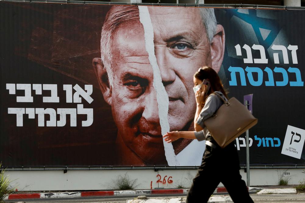 A woman walks past a Blue and White party election campaign banner depicting its leader, Israeli Defense Minister Benny Gantz, alongside Israeli Prime Minister Benjamin Netanyahu ahead of the March 23 ballot, in Tel Aviv, Israel, March 14, 2021. REUTERS/Corinna Kern/File Photo