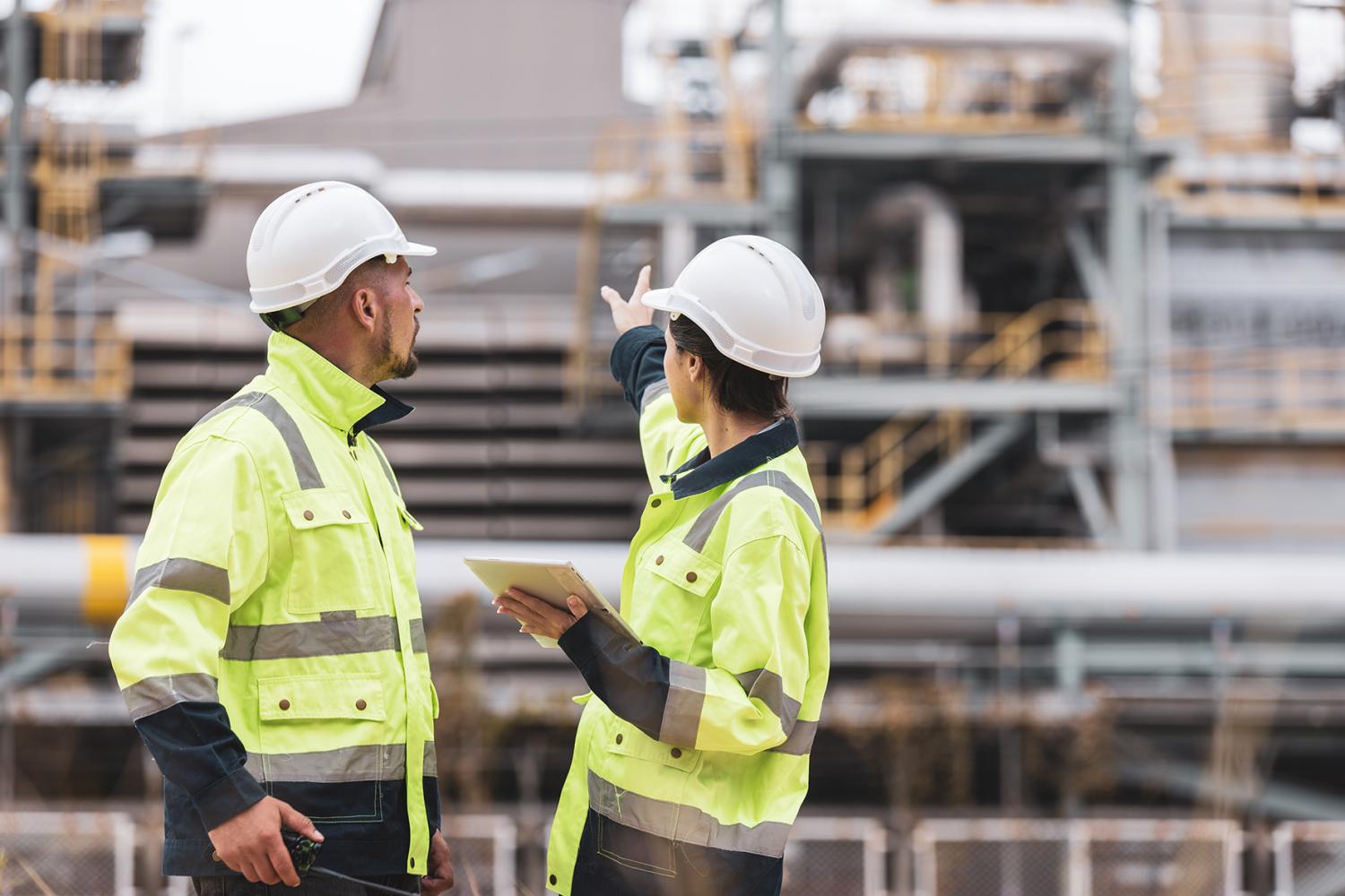 group of industrial engineers workers in a refinery - oil and gas processing equipment and machinery, engineers collaborate with a laptop, blueprint, and digital tablet at the oil storage tanks site.