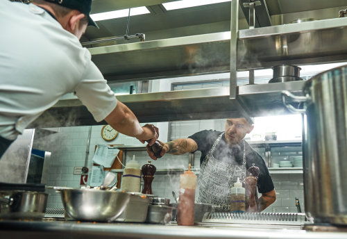 Two workers in food preparation jobs help each other by passing a pepper grinder